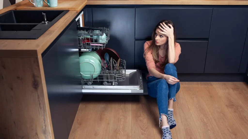 Frustrated homeowner sitting on the kitchen floor beside an open dishwasher