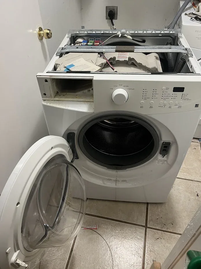 White front-load washer with its door and top panel open during a repair